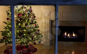 Outside window view of lighten Christmas tree and glowing fireplace during night time in family living room home 