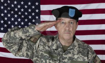 Mature man saluting while wearing military outer shirt plus beret with US flag in background 