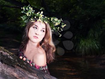Beautiful girl in national dress and wreath stand in the forest near river.