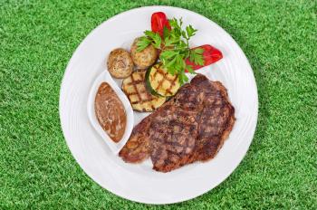 Grilled steak, baked potatoes and vegetables on white plate on green grass. Closeup.