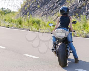 Young beautiful blonde girl in trendy blue jeans and a black t-shirt rides on modern motorcycle.  Outdoor portrait in soft sunny colors.
