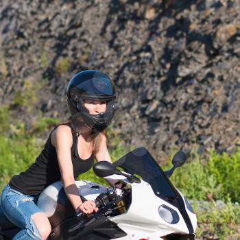 Young beautiful blonde girl in trendy blue jeans and a black T-shirt on the nature behind the wheel of modern motorcycle. Portrait in soft sunny colors.