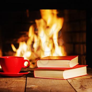 Red cup of tea or coffee and old books near fireplace on wooden table. Winter and Christmas holiday concept.
