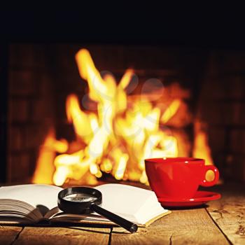 Red cup of coffee or tea, magnifier glass and old book on wooden table near  fireplace. Winter and Christmas holiday concept. Photo with retro filter effect.