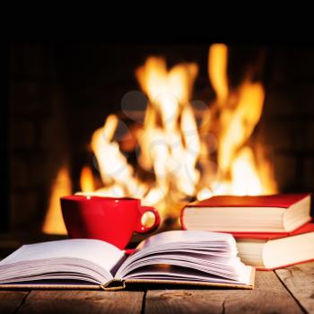Red cup of coffee or tea and old books on wooden table near  fireplace. Winter and Christmas holiday concept.

