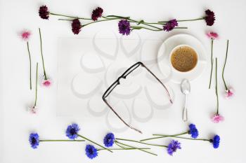 Paper, cup of coffee and glasses with wreath frame from petals of wildflowers on white background. Overhead view. Flat lay.