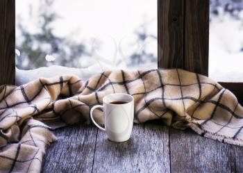White cup of coffee or tea and woolen plaid located on stylized wooden window sill. Winter concept of comfort and relaxation.