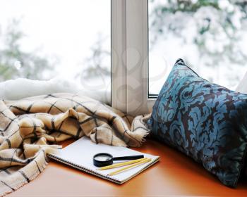 Open notepad, magnifier glass, pillow, pencils and beige warm plaid located on stylized wooden windowsill. Winter concept of comfort and relaxation.