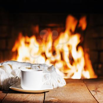 White cup of tea or coffee and woolen scarf near fireplace on wooden table. Winter and Christmas holiday concept.