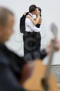 A young loving couple embrace while a street musician plays his guitar (out of focus) in the foreground