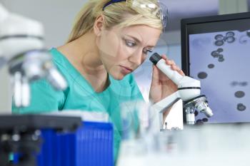 A blond female medical or scientific researcher or woman doctor using her microscope in a laboratory with a computer monitor next to her.