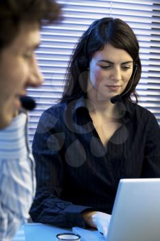 Two young business people sitting at desk, having a teleconference, talking on headset.