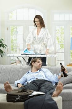 Tired businessman resting on couch with beer in hand. Angry wife ironing in the background.