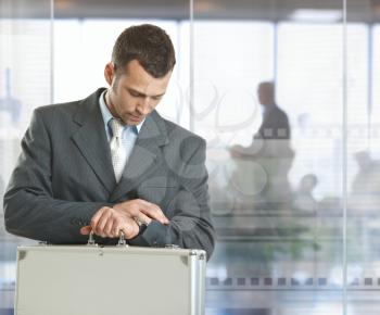 Businessman standing in office lobby looking at his watch, checking time.