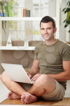 Casual man using laptop computer at home sitting on floor in living room, smiling.