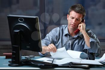 Mid-adult businessman speaking on landline phone working overtime in office checking papers.