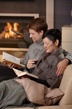Young couple hugging on sofa in front of fireplace at home, reading books, smiling.
