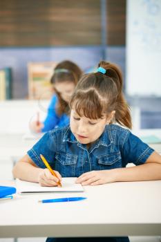 Young girl writing at school sitting in class with other girl in background.