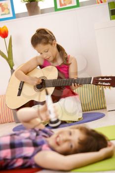 Young girl in focus playing guitar, other girl singing into microphone.