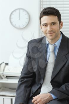 Portrait of yuong handsome businessman wearing grey suit and blue shirt sittigng on desk at office, smiling.