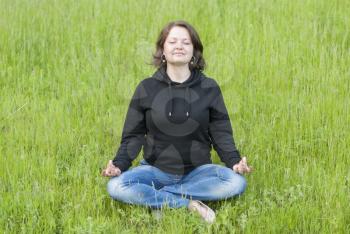 Beautiful young girl meditating on the green grass in the park.