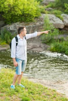Tourist standing near a mountain river pointing thumb into the distance.
