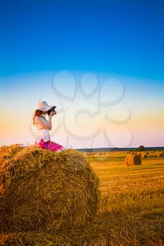 Beautiful Young Girl Woman In Dress Sitting On Haystack And Making Photos With Vintage Film Camera In Sunny Day In Field, Meadow On Sunrise, Sunset