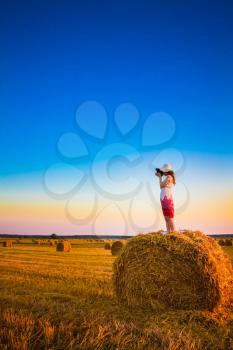Beautiful Young Girl Woman In Dress Staying On Haystack And Making Photos With Vintage Film Camera In Sunny Day In Field, Meadow On Sunrise, Sunset