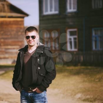 Young Handsome Man Staying Near Old Wooden House In Autumn Day, Relaxing,  Thinking, Smiling. Casual Style - Jeans, Jacket, Sunglasses