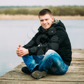 Young Handsome Man Sitting On Wooden Pier In Autumn Day, Relaxing,  Thinking, Smiling. Casual Style - Jeans, Jacket