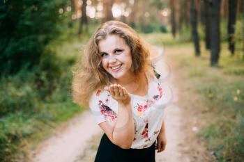 Portrait Of Young Pretty Plus Size Caucasian Happy Smiling Girl Woman With Blue Eyes, Wavy Brown Long Hair In White T-Shirt With Floral Print Sending A Kiss To Camera, Summer Forest Background.
