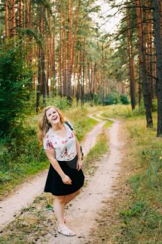 Young Pretty Plus Size Caucasian Happy Smiling Laughing Girl Woman With Wavy Brown Long Hair In White T-Shirt And Black Short Skirt Standing Full-Length On Road In Summer Pine Forest