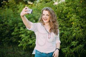 Young Beautiful Pretty Plus Size Caucasian Girl Woman Dressed In White Blouse Enjoying Life, Smiling, Having Fun And Taking Selfie Photo On Smartphone In Summer Green Forest Park Bushes Background