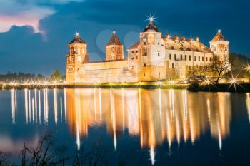 Mir, Belarus. Picturesque View Of Mir Castle Complex In Bright Evening Illumination With Glow Reflexions On Lake Water. Famous Landmark, Ancient Gothic Monument Of Feudalism Under Blue Dramatic Sky.