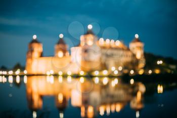 Mir, Belarus. Blurred Boke Bokeh Focus Of Mir Castle Complex In Night Illumination And Reflexions On Lake Water Surface In Warm Yellow Colors.