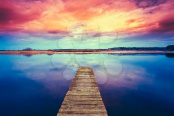 Colorful sunset dramatic sky over wooden boards pier on Calm Water Of Lake, River. Nature Background.