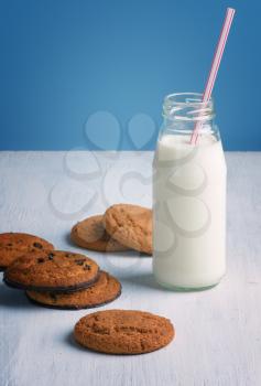 Chocolate chip cookies with a milk bottle with a straw on a white wooden table and blue background. 