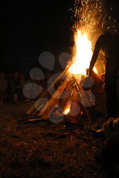 Blurred People sit at night around a bright bonfire and silhouette of a man throwing firewood into a fire