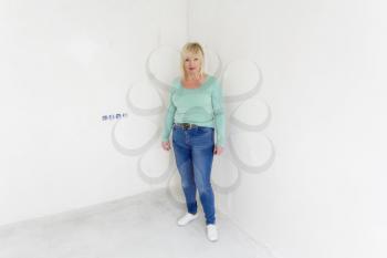 Blond woman in green and blue jeans stand inside empty room
