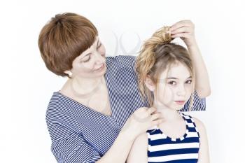 Mother is making hairstyle to her daughter near white wall in striped clothes