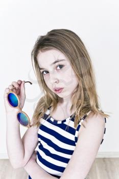 Blond girl eleven years old standing near white wall with green sunglasses