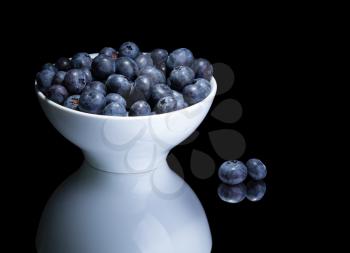 Blueberries in white bowl on a mirror. Isolated on black.