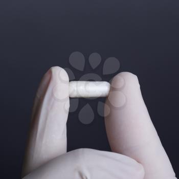 Hand in white latex glove holding white capsule on dark background
