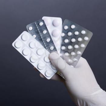 Male hand in glove holding pills in blisters on dark background. Square format.