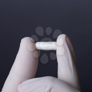 Hand in white latex glove holding white capsule. Doctor or nurse showing medicine. Dark background