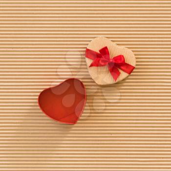 Heart shaped gift box with red ribbon and bow on corrugated cardboard background. Square format.