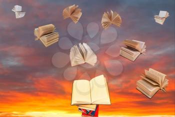 open book above of stack of books and sunset sky with flying books background