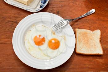 breakfast with two fried eggs on white plate, fresh toast and dairy butter in butterdish on wooden table