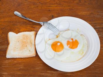 breakfast with two fried eggs on white plate and toast on wooden table