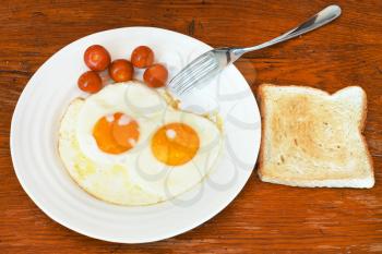 breakfast with two fried eggs on white plate, cherry tomatoes, fresh toast on wooden table